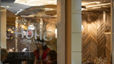Restaurant dining area seen through a glass window with repeated 'HOME' word art; warm lighting and tables set for dinner.